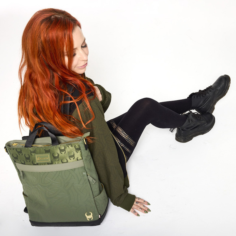 Red-headed woman sitting on the floor wearing the Loungefly COLLECTIV Marvel Loki The WEEKENDR Hooded Jacket and black leggings, leaning back against the Loungefly COLLECTIV Marvel Loki The CREATIV Convertible Tote Bag